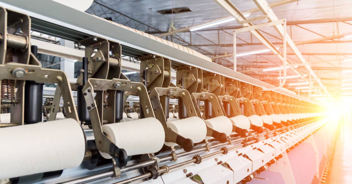 Textile manufacturing machines processing multiple large spools of white thread inside an industrial warehouse.